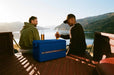 Two people enjoying drinks by a lake with a blue cooler box in the foreground.
