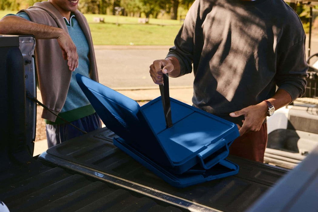 A man standing next to a van, collapsing a portable coolbox by folding its soft sides inward on the vehicle’s loading area, preparing it for compact storage.