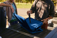 A man standing next to a van, collapsing a portable coolbox by folding its soft sides inward on the vehicle’s loading area, preparing it for compact storage.